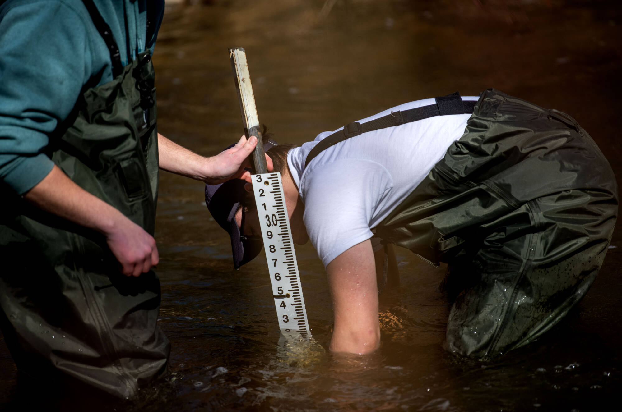 Two students standing in a stream with waders on measuring the depth of the stream